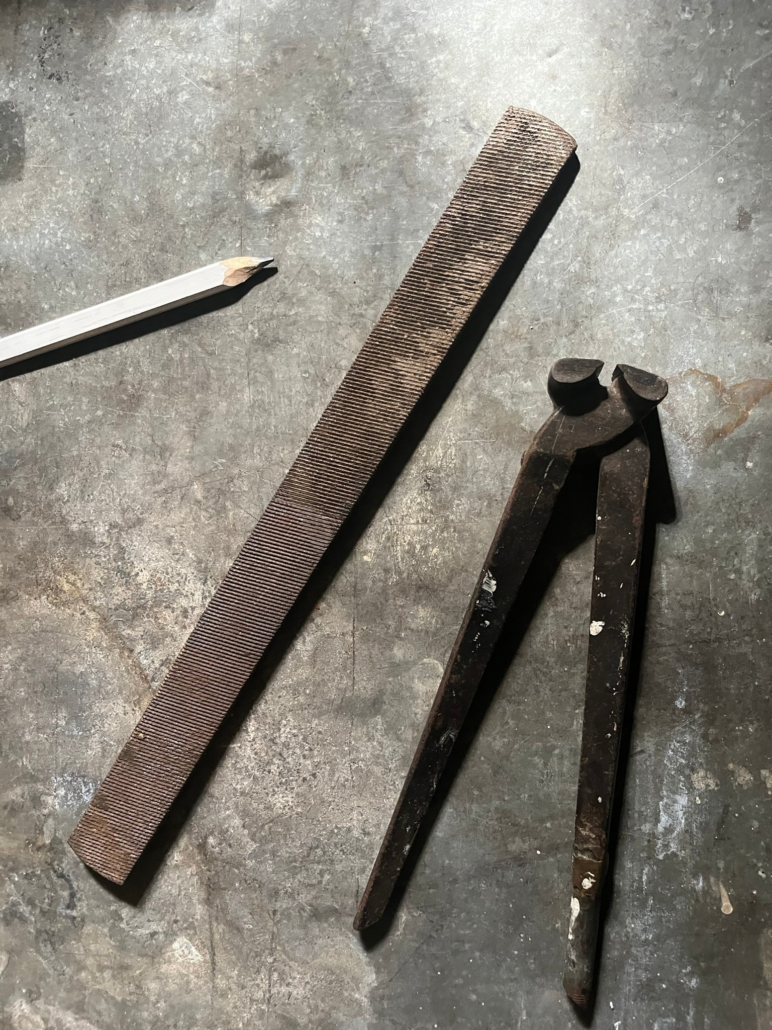 An enormous coarse file next to a pair of heavy pincers, with a carpenter's pencil for scale