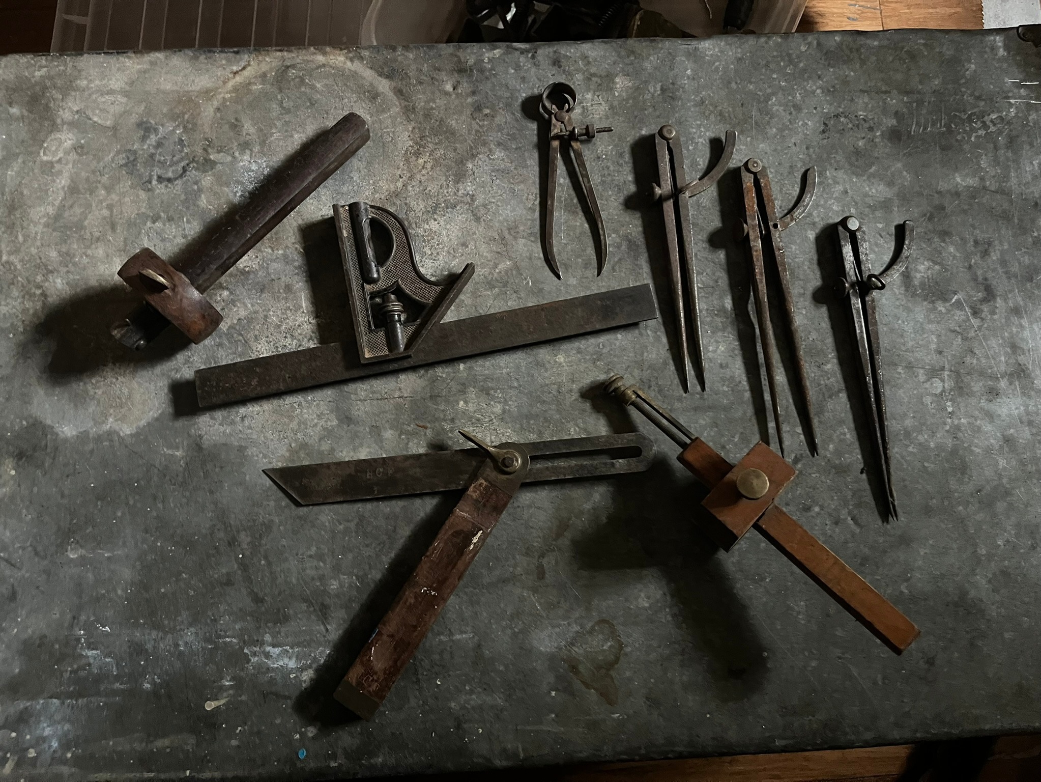 A combination square, sliding bevel, marking gauge, and several pairs of dividers and calipers laid out on the bench