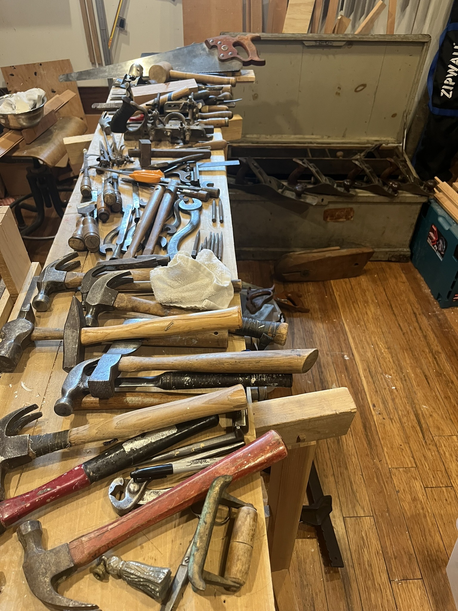 A workbench covered end to end with old hammers, saws, planes, and hand tools, with the open chest behind it